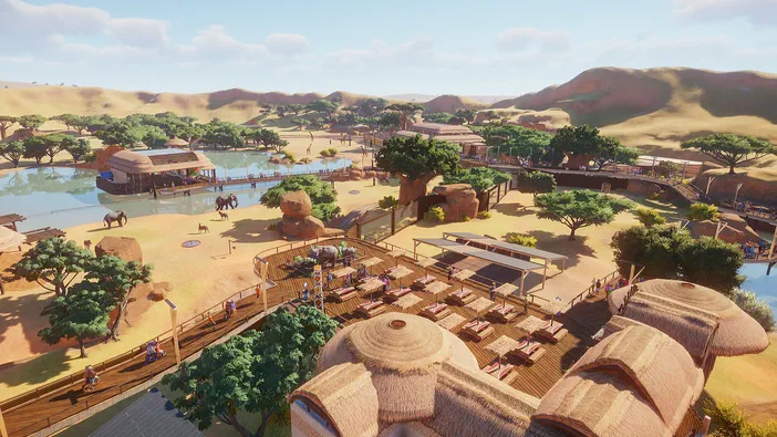 Capture d'écran du jeu Planet Zoo montrant un zoo en plein air avec des éléphants dans une étendue d'eau et sur le sable, des visiteurs marchant sur des sentiers boisés entourés de bâtiments à toit de chaume au milieu d'un paysage désertique.
