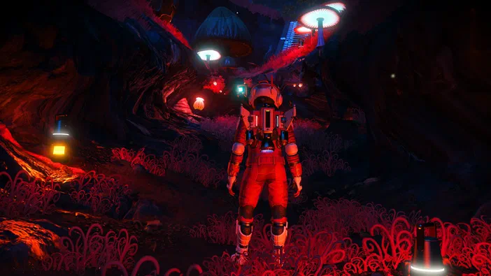 Dans le jeu vidéo No Man's Sky, un astronaute en combinaison rouge se tient dans une grotte aux parois rocheuses sombres et à la végétation bioluminescente émettant des lueurs rouges.