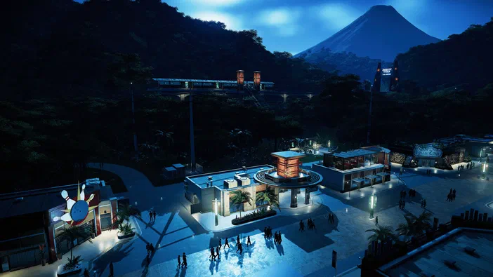 Capture d'écran du jeu vidéo Jurassic World Evolution montrant une vue nocturne d'un parc de dinosaures avec des bâtiments illuminés, un bowling et des montagnes au loin.