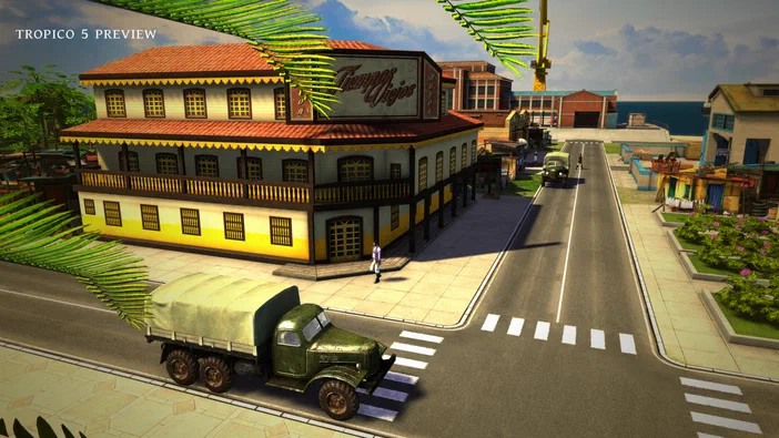 Capture d'écran de Tropico 5 montrant une rue avec un grand bâtiment jaune au toit rouge, des camions verts sur la route et de la végétation tropicale en encadrement.