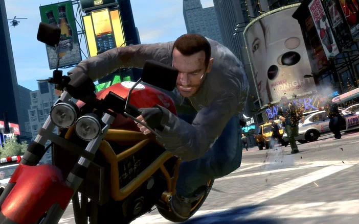 Un homme est penché en avant sur une moto rouge au milieu d'une rue urbaine ensoleillée avec des immeubles et publicités en arrière-plan dans le jeu Grand Theft Auto IV.