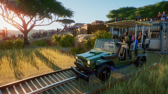 Screenshot de Planet Zoo montrant un véhicule vert type Jeep modifié circulant sur une voie ferrée dans le parc animalier simulé, avec des visiteurs en arrière-plan.