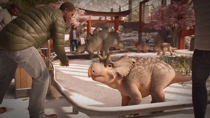 Dans le jeu vidéo Jurassic World Evolution 3, un homme nourrit un triceratops dans un parc de dinosaures où d'autres dinos et visiteurs sont visibles en arrière-plan sous des arbres en fleurs.