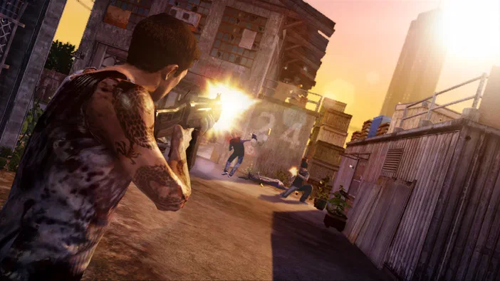 Scène d'action du jeu Sleeping Dogs où un personnage avec tatouages tire son fusil à pompe créant une explosion lumineuse jaune, tandis que deux autres personnages sont visibles en arrière-plan dans une rue urbaine aux bâtiments délabrés sous un ciel orange.