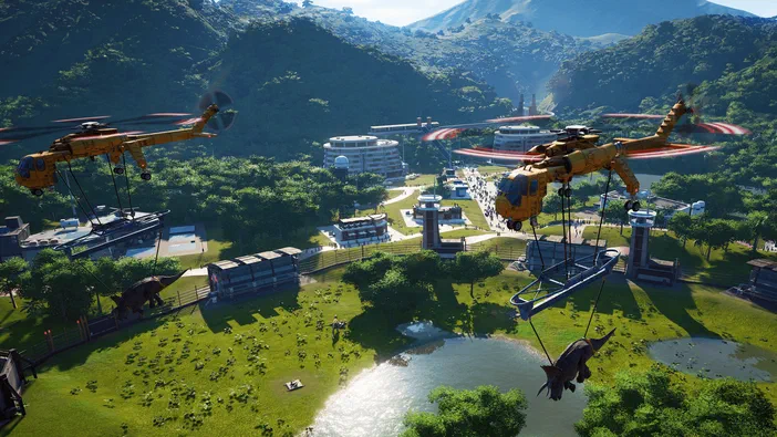 Capture d\'écran du jeu Jurassic World Evolution montrant deux hélicoptères transportant des dinosaures au-dessus d\'une base entourée de bâtiments et de montagnes.