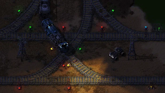 Vue aérienne d'une base Factorio avec des rails complexes, plusieurs trains et un véhicule blindé sur un terrain désertique éclairé par des lumières colorées.