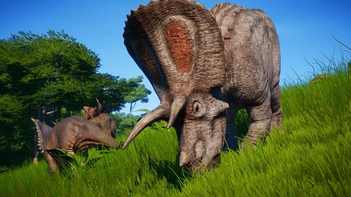Capture d'écran du jeu Jurassic World Evolution montrant deux dinosaures triceratops dans un environnement herbeux sous un ciel bleu.