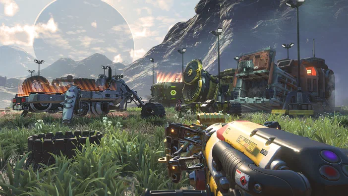 Capture d'écran de No Man's Sky : arme tenue par le joueur au premier plan, véhicules futuristes stationnés dans un champ herbeux devant des montagnes et une planète en arrière-plan.