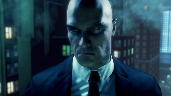 Un homme chauve vêtu d'un costume sombre et d'une cravate noire regarde intensément dans cette capture d'écran du jeu vidéo Hitman: Absolution, avec un fond de ville illuminée la nuit.