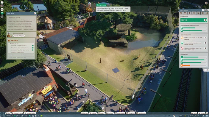 Capture d'écran du jeu Planet Zoo montrant une vue en hauteur d'un zoo avec un enclos pour lions, des visiteurs sur les sentiers et des bâtiments, accompagnée d'interfaces utilisateur affichant les objectifs de mission et le statut d'un invité nommé Rudolf Ragland à droite.