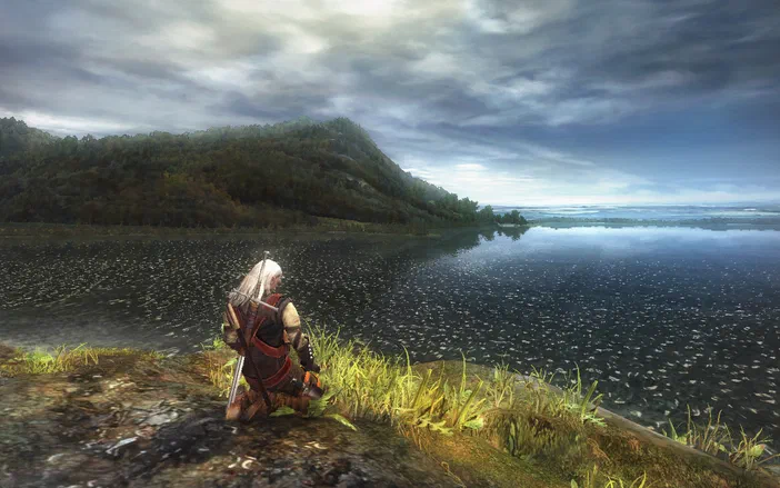 Dans The Witcher: Enhanced Edition, un personnage aux cheveux blancs est assis sur la berge d'un lac entouré de collines boisées sous un ciel nuageux.