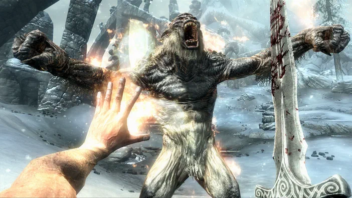 Dans The Elder Scrolls V: Skyrim, un personnage de premier plan tend la main vers une créature monstrueuse qui hurle dans un paysage enneigé.