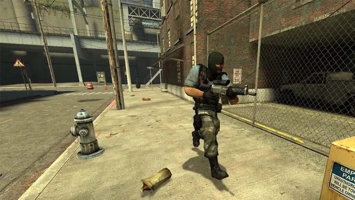 Dans Counter-Strike: Source, un terroriste en tenue de camouflage et masque noir court sur le trottoir d'une rue urbaine tout en tenant une arme à feu.
