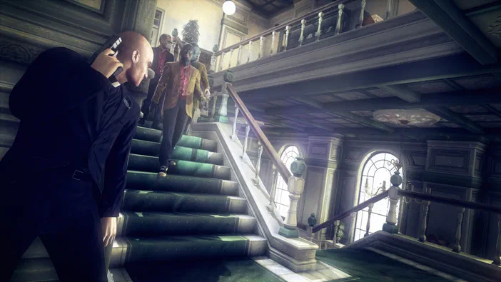 Dans Hitman: Absolution, un assassin en costume noir se tient sur une grande escalier vert à l'intérieur d'un bâtiment luxueux. Il vise avec son arme alors que plusieurs cibles armées descendent les marches vers lui.