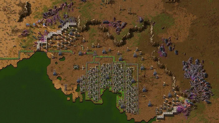 Capture d'écran du jeu Factorio montrant une base industrielle complexe avec des bâtiments et des unités sur un terrain marron bordé d'eau verte.
