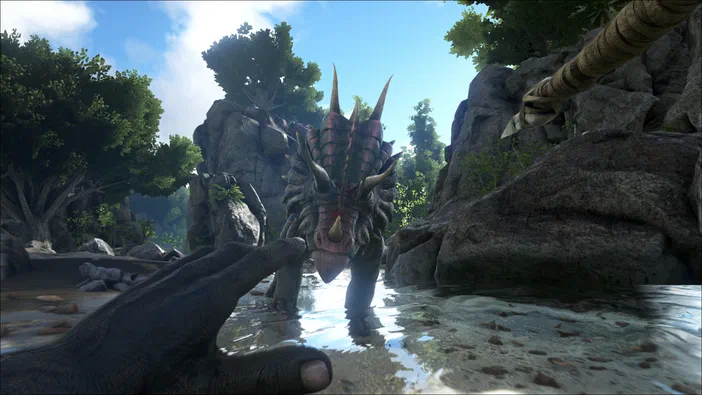 Capture d'écran du jeu vidéo ARK: Survival Evolved montrant une main humaine tendue vers un dinosaure à crête rouge se tenant dans l'eau, avec des rochers et de la végétation en arrière-plan.