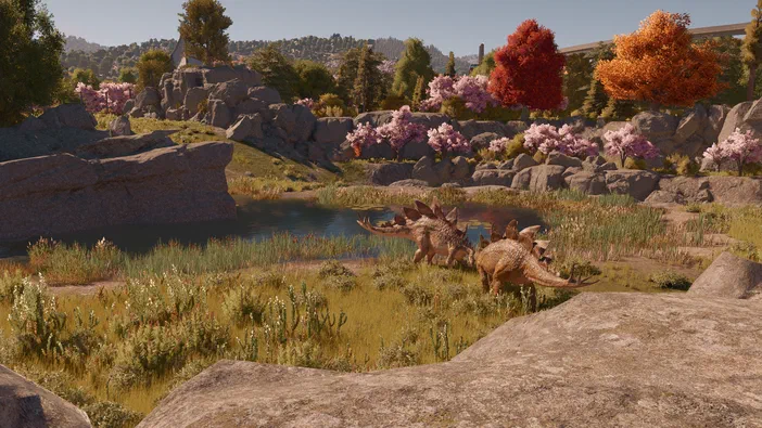 Deux stégosaures se tiennent près d'un étang dans un paysage rocheux parsemé de fleurs et d'arbres aux couleurs automnales, scène issue du jeu vidéo Jurassic World Evolution.