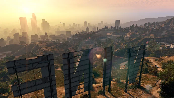 Vue aérienne du jeu vidéo Grand Theft Auto V Enhanced montrant les lettres géantes 'F' et 'N' sur une colline avec un paysage urbain au loin sous un soleil couchant.