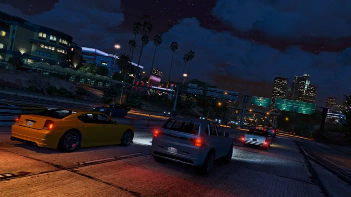 Capture d'écran de Grand Theft Auto V Enhanced montrant trois voitures sur une autoroute nocturne avec des panneaux indiquant Los Santos et Downtown.