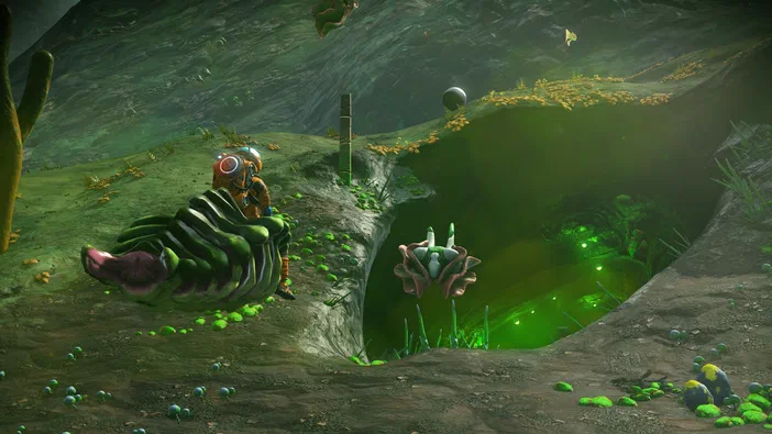 Capture d'écran du jeu vidéo No Man's Sky montrant un personnage en combinaison spatiale sur une planète aux couleurs vertes et brunes avec des formations rocheuses étranges.