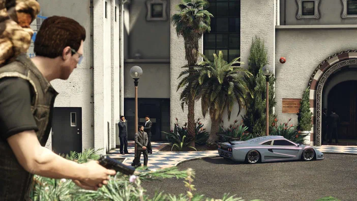 Dans Grand Theft Auto V Legacy, un personnage tient une arme devant des hommes en costume et une voiture de sport dans la cour d'un bâtiment.