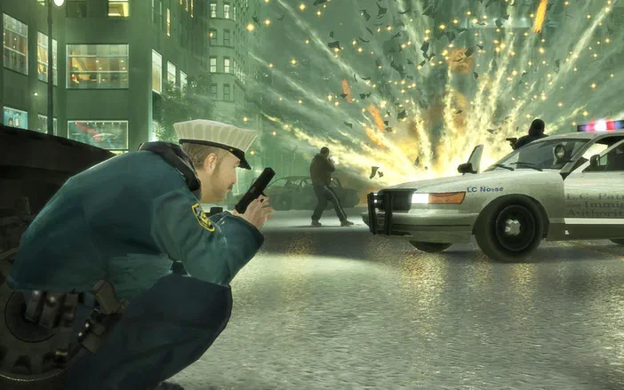 Dans Grand Theft Auto IV, un policier en tenue bleue tire son arme au premier plan tandis qu'une voiture de police et des explosions illuminent la rue urbaine.