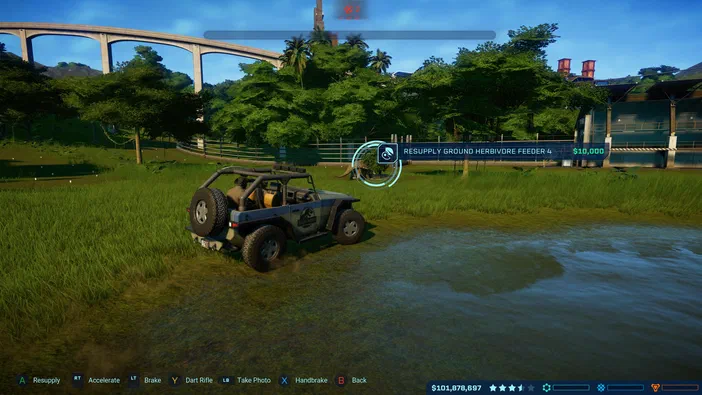 Dans Jurassic World Evolution, une jeep est garée près d'un étang dans un parc de dinosaures avec des arbres et un pont en arrière-plan.