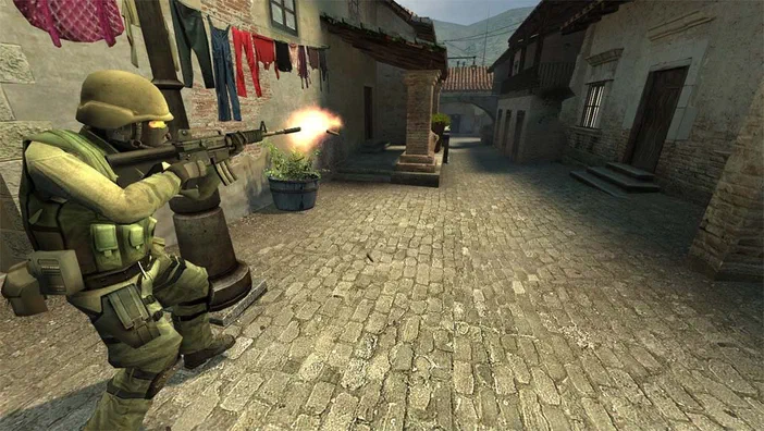 Dans Counter-Strike: Source, un soldat en tenue de camouflage tire son fusil d'assaut dans une ruelle pavée bordée de bâtiments et de linge séchant.