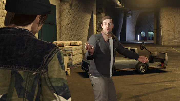 Dans Grand Theft Auto V Legacy, un personnage masculin en veste grise s'adresse à une autre personne dans un entrepôt souterrain avec des palettes et un chariot.