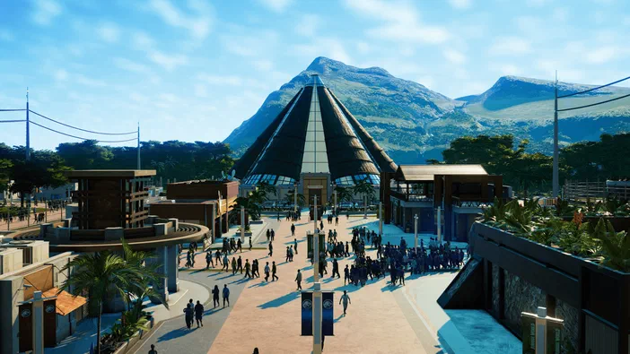 Vue d'un parc de dinosaures dans le jeu Jurassic World Evolution avec des bâtiments et une montagne en arrière-plan.