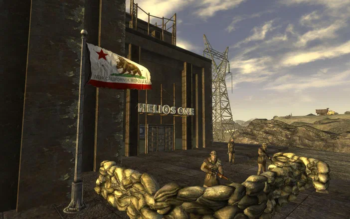 Capture d'écran du jeu Fallout: New Vegas montrant l'entrée de la base Helios One avec des soldats postés derrière un rempart de sacs de sable et le drapeau de la République Nouvelle-Californie flottant au vent.