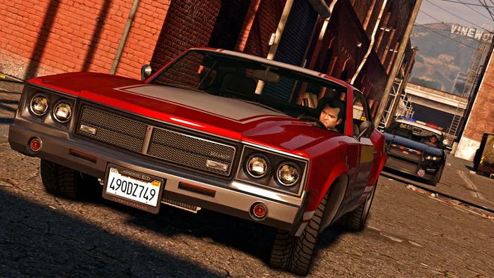 Grand Theft Auto V Enhanced : une voiture rouge conduite par un homme dans une rue urbaine avec des bâtiments en briques et une police derrière.
