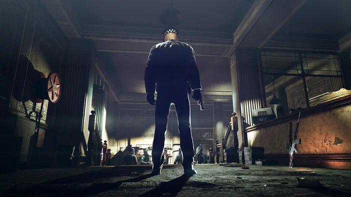 Capture d'écran du jeu vidéo Hitman: Absolution montrant l'assassin de dos dans une pièce sombre avec des armes au sol.