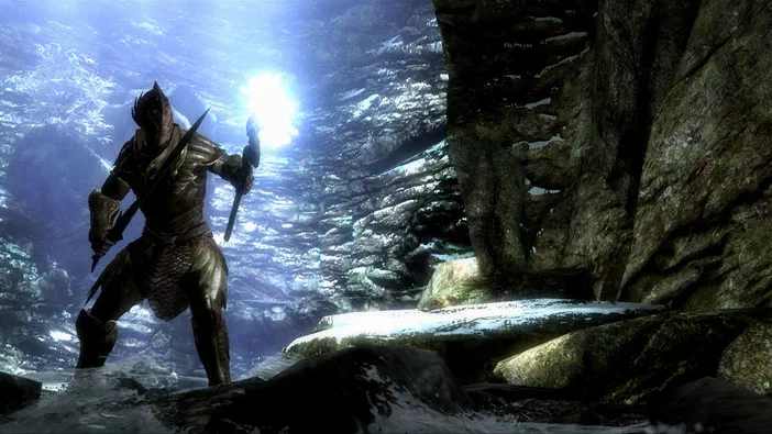 Dans The Elder Scrolls V: Skyrim, un personnage en armure se tient dans une grotte enneigée sous la lumière du jour.