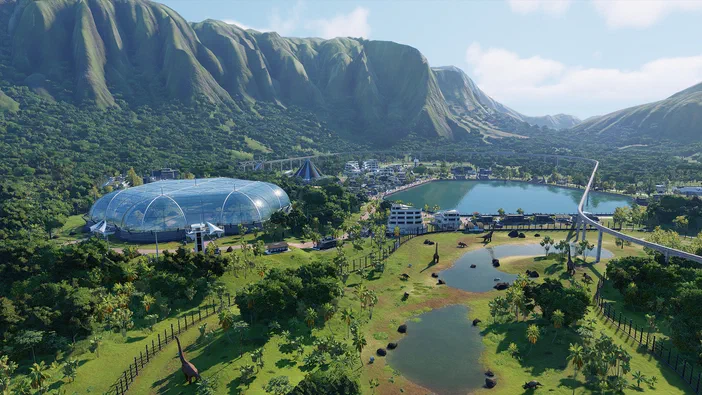 Vue aérienne d'un parc de dinosaures dans le jeu Jurassic World Evolution 2 avec des bâtiments modernes et une montagne en arrière-plan.