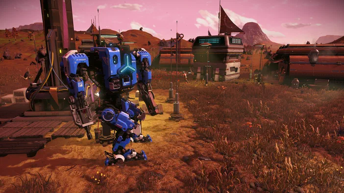 Dans le jeu No Man's Sky, un robot bleu se tient au premier plan devant une base avec des véhicules et structures dans un paysage aride sous un ciel rose.