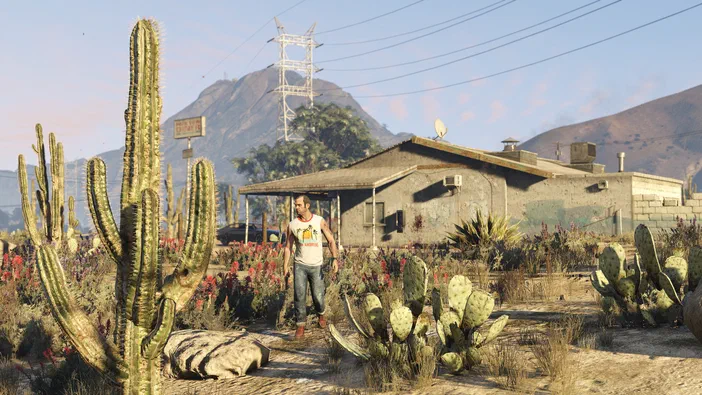 Dans Grand Theft Auto V Legacy, un personnage masculin marche dans une zone désertique parsemée de cactus devant une maison en béton.