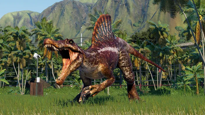 Capture d'écran du jeu Jurassic World Evolution 2 montrant un Spinosaurus en pleine course dans une savane verdoyante avec des palmiers et des montagnes à l'arrière-plan.