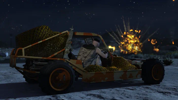 Dans Grand Theft Auto V Legacy, un personnage est assis dans une dune orange et beige surchargée de filets de camouflage. En arrière-plan nocturne sous les étoiles, des explosions éclatent au loin.