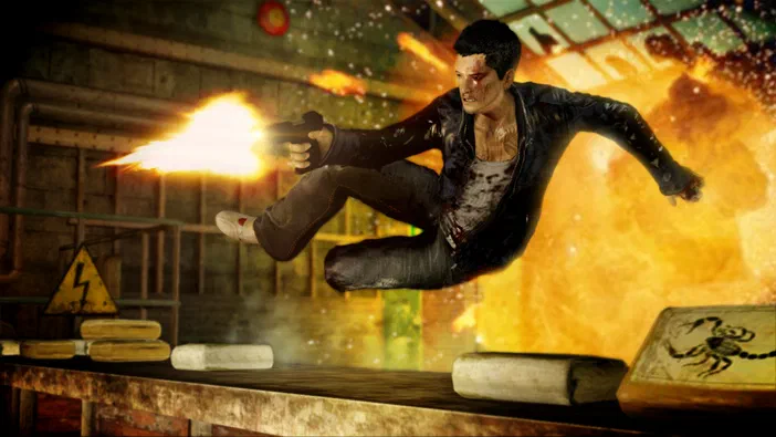 Dans le jeu Sleeping Dogs™, un personnage masculin en tenue de combat saute au-dessus d'une table tout en tirant avec une arme à feu dans un environnement industriel explosif.