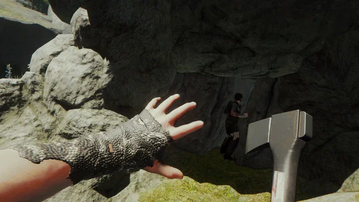 Dans le jeu vidéo The Forest, une main gantée tendue vers l'avant et un personnage en arrière-plan dans une grotte rocheuse.
