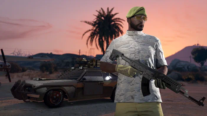 Dans Grand Theft Auto V, un personnage masculin vêtu d'un uniforme de camouflage et coiffé d'une casquette verte tient une arme à feu. Derrière lui se trouve une voiture modifiée dans un décor désertique au coucher du soleil.