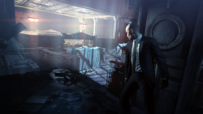 Dans Hitman: Absolution, l'agent 47 en costume observe une pièce désordonnée et éclairée par des lampes suspendues.