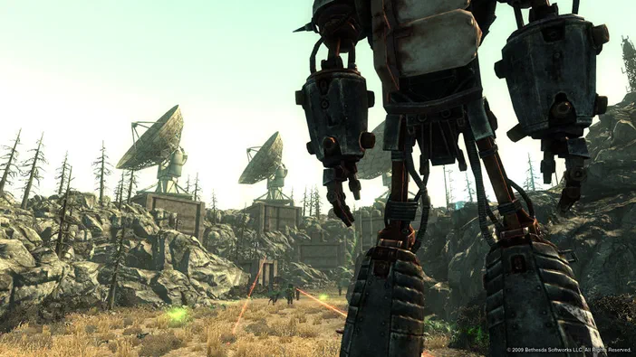 Capture d'écran de Fallout 3 montrant un robot imposant au premier plan et des antennes paraboliques sur une colline aride en arrière-plan.
