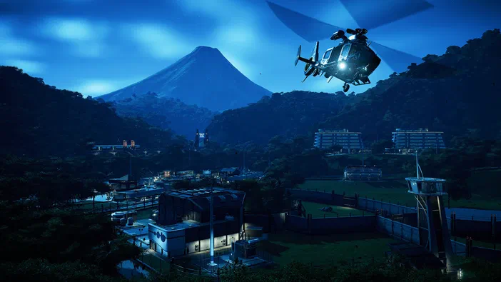 Vue noctuelle d'un parc de dinosaures avec une montagne en arrière-plan et un hélicoptère survolant la zone dans le jeu Jurassic World Evolution.