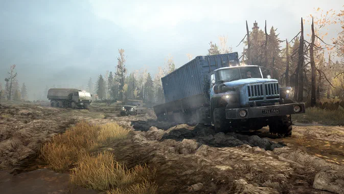 Dans le jeu MudRunner, un camion bleu tractant une benne bleue s'engage dans la boue d'un terrain forestier avec deux autres véhicules en arrière-plan.