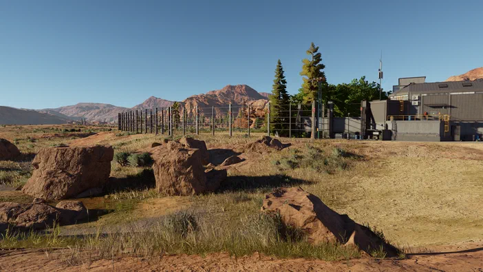 Vue d'un parc de Jurassic World Evolution 3 avec des rochers, une clôture et un bâtiment dans un paysage aride sous un ciel bleu.