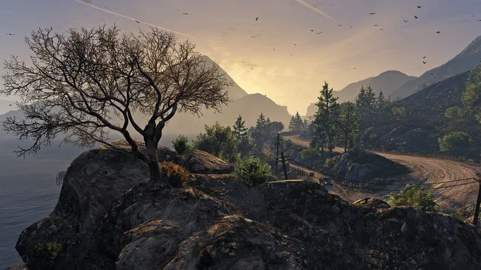 Vue de Grand Theft Auto V Legacy montrant un paysage montagneux au coucher du soleil avec une route sinueuse bordée d'arbres et des oiseaux en vol.