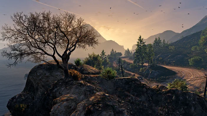Capture d'écran de Grand Theft Auto V Enhanced montrant un paysage montagneux au coucher du soleil avec une route sinueuse et des arbres.