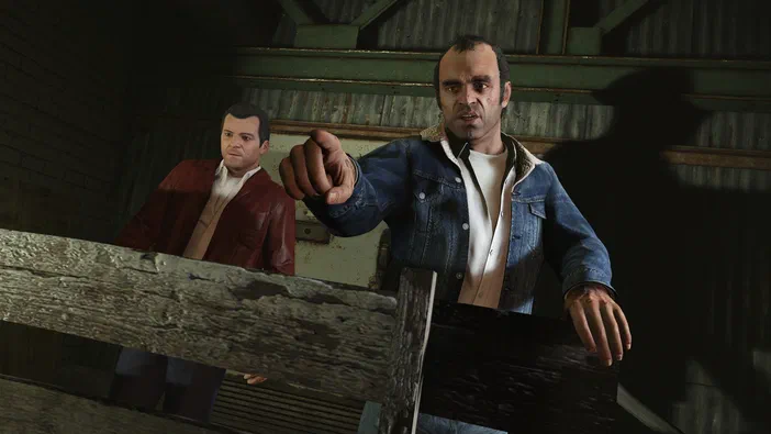 Dans Grand Theft Auto V Legacy, un homme en jean pointe vers l'avant tandis qu'un autre personnage en veste rouge se tient derrière lui dans une structure de bois.